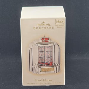 May include: A Hallmark Keepsake Christmas ornament in a box. The ornament is a silver jukebox with red and white details. The box reads "Santa's Jukebox" and "christmas ornament - décoration de noël". The ornament has the words "Magic Light, Sound, and Movement".