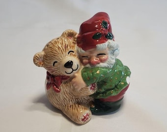 May include: A ceramic Christmas figurine of a gnome hugging a teddy bear. The gnome wears a red hat with holly, a green jacket with gold dots, and red pants. The teddy bear is tan with a red bow.