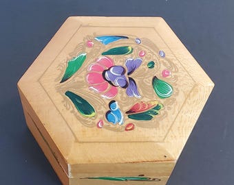 May include: Hexagonal wooden box with a floral design. The box is light brown with a painted floral pattern in pink, purple, blue, and green. The box is closed and sits on a dark surface.
