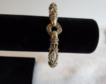 May include: Silver and gold-toned bracelet with intricate detailing. The bracelet showcases a series of decorative links, including diamond-shaped and cylindrical elements. The design incorporates detailed patterns and textures, creating a vintage aesthetic.