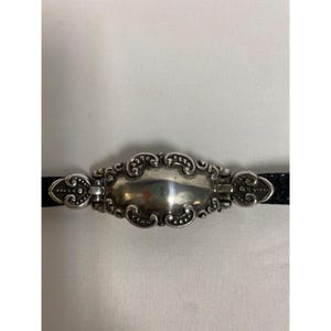 Op de afbeelding: Een zilverkleurige metalen armband met een sierlijk, ovaalvormig middelpunt. Het middelpunt wordt geflankeerd door decoratieve elementen en bevestigd aan een zwarte band. Het ontwerp heeft ingewikkelde krullen en kralen details.