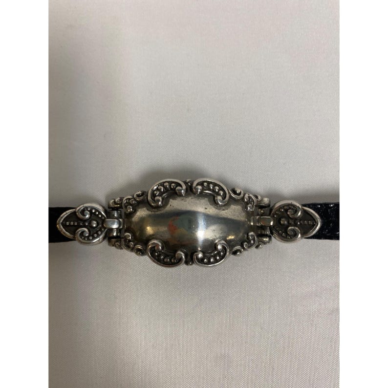 Op de afbeelding: Een zilverkleurige metalen armband met een sierlijk, ovaalvormig middelpunt. Het middelpunt wordt geflankeerd door decoratieve elementen en bevestigd aan een zwarte band. Het ontwerp heeft ingewikkelde krullen en kralen details.