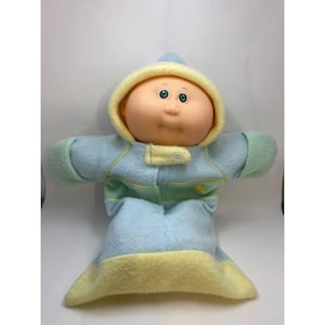 May include: A Cabbage Patch Kids doll in a blue, yellow, and green hooded jumpsuit. The doll has a peach-coloured face with green eyes and a small nose. The jumpsuit has a button closure and a yellow hood lining.
