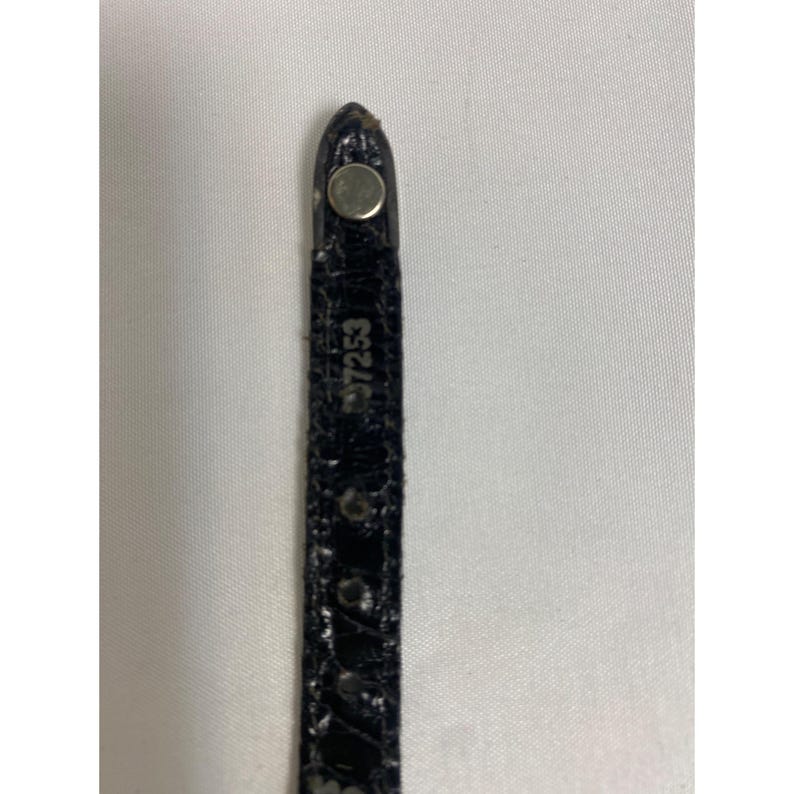 Op de afbeelding: Close-up van een zwart horlogebandje met een zilveren metalen sluiting. De band heeft een textuur, krokodilachtig patroon. Het nummer "97253" staat op de band.