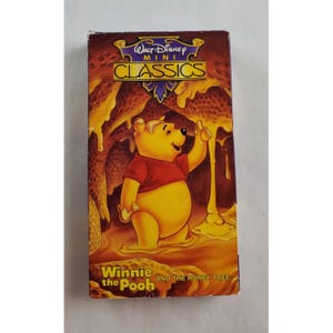 May include: A VHS tape of "Winnie the Pooh and the Honey Tree" from the Walt Disney Mini Classics series. The cover features Winnie the Pooh in a red shirt, reaching for honey. The background shows honey dripping from a tree.