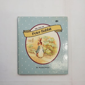 Puede incluir: Una copia vintage de "The Tale of Peter Rabbit" de Beatrix Potter. La portada del libro es azul claro con un patrón blanco repetido. El título está en una pancarta dorada sobre una ilustración circular de Peter Rabbit en un jardín.