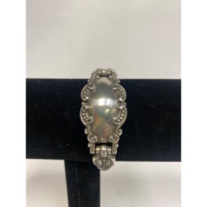 Op de afbeelding: Zilverkleurige armband met een sierlijk, vintage ontwerp. De armband heeft een centrale ovale plaat met decoratieve randen en verbindingsschakels. Het metaal heeft een gepolijste afwerking, die licht reflecteert.