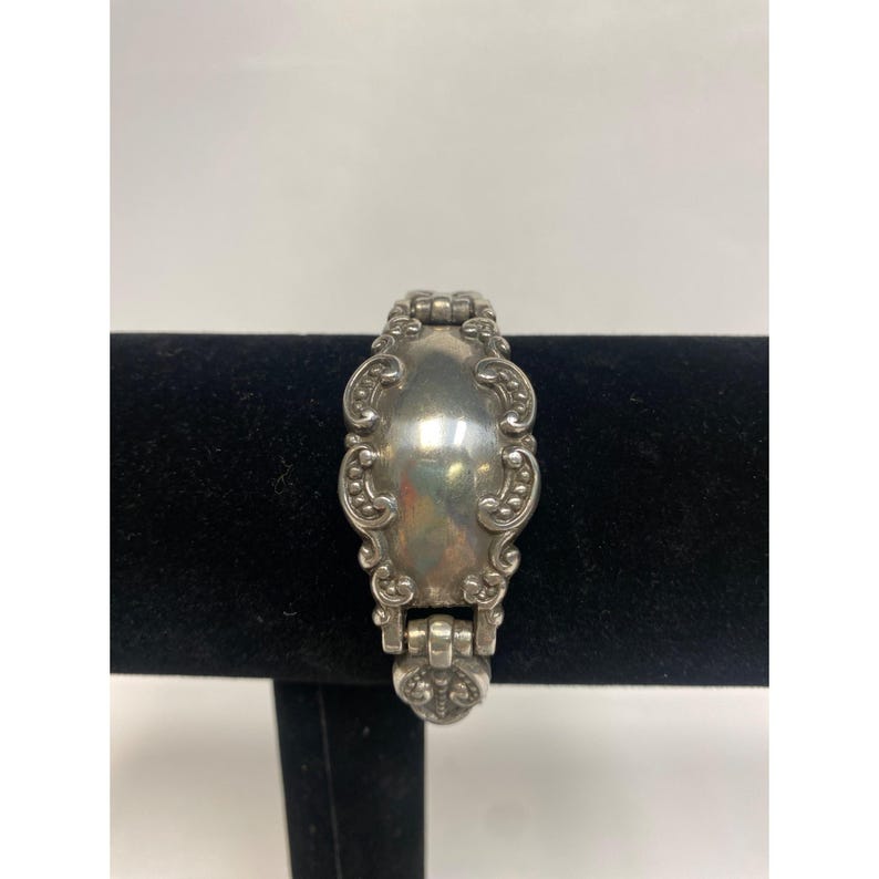 Op de afbeelding: Zilverkleurige armband met een sierlijk, vintage ontwerp. De armband heeft een centrale ovale plaat met decoratieve randen en verbindingsschakels. Het metaal heeft een gepolijste afwerking, die licht reflecteert.