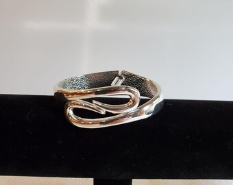 May include: A silver bracelet with a distinctive, flowing design. The bracelet has a hinged clasp and a polished, curved pattern. The design includes two looping elements, creating an elegant and modern look. The bracelet is displayed on a black surface.