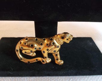 May include: Gold-coloured metal leopard brooch with black spots and a detailed collar. The brooch is displayed on a black velvet surface, showcasing its intricate design. A stylish accessory.
