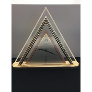May include: A gold-toned Seiko quartz clock in a triangular shape. The clock face is grey with black hands and the numbers 12 and 6. The clock is encased in clear acrylic and gold-toned metal.