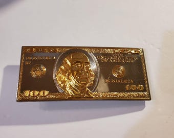 May include: A gold-coloured metal belt buckle shaped like a £100 bill. The buckle features a portrait of Benjamin Franklin, the words "UNITED STATES OF AMERICA," and the number "100."