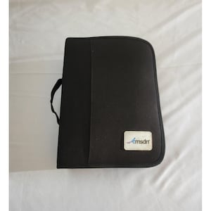 May include: Black zippered portfolio with a handle. The portfolio has a rectangular shape with rounded corners. A small white label with the text "msdn" is on the lower right corner. The portfolio is closed.