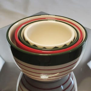 Op de afbeelding: Een set van vier in elkaar geneste keramische mengkommen. De grootste kom is donkergroen met rode en witte strepen. De andere kommen zijn rood, groen en crèmekleurig, met een decoratieve rand. De kommen zijn in elkaar gestapeld.