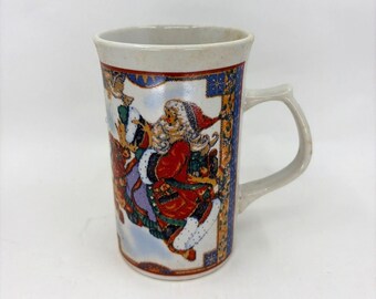 May include: A ceramic Christmas mug with a cream-colored rim and handle. The mug features a colorful illustration of Santa Claus riding a reindeer, with a blue and gold border. The mug is approximately 4 inches tall.