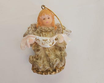 May include: A small angel figurine with golden hair and wings. The angel is wearing a gold and cream-coloured dress and holding a banner that reads "Joy". A gold string is attached for hanging.