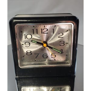 May include: A black Quartzmatic Westclox travel alarm clock with a silver face. The clock has a rectangular shape with a silver dial, black hands, and large, easy-to-read numbers. The clock face is protected by a clear cover.
