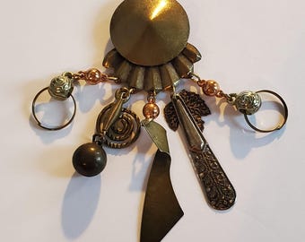 May include: A vintage-style brooch with a central, circular, bronze-colored element. It features a cone-shaped top and a ruffled base. Dangling from the base are various charms, including a spiral, a leaf, a ball, and two rings, all in shades of bronze and copper.