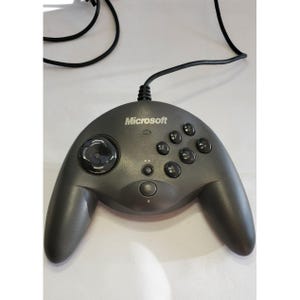 May include: A gray Microsoft brand game controller with a black cable. The controller has a circular joystick, several black buttons, and the Microsoft logo. The controller is designed with two handles.