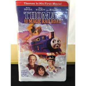 Puede incluir: Cinta VHS de la película "Thomas y el Ferrocarril Mágico". La portada presenta una locomotora azul con una expresión de sorpresa, el título de la película y los actores Peter Fonda, Mara Wilson y Alec Baldwin. También se ve el texto "Little Engines CAN Do Big Things...".
