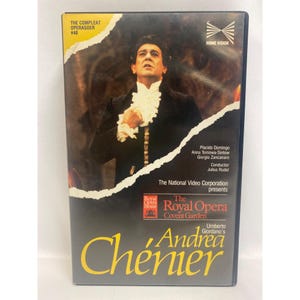 Pode incluir: Uma fita VHS da ópera "Andrea Chénier" com Plácido Domingo. A capa tem um fundo escuro com uma foto de um homem em roupa formal. O texto inclui "The Royal Opera Covent Garden" e "Home Vision."