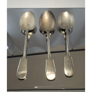 May include: Three antique silver spoons with oval bowls and long, rectangular handles. The spoons are arranged vertically, reflecting on a mirrored surface. The spoons are likely for serving or eating.