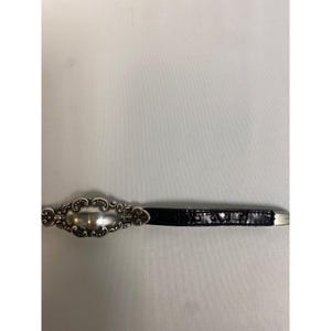 Op de afbeelding: Een zilverkleurige gesp met een sierlijk, ovaal ontwerp is bevestigd aan een zwarte, getextureerde riem. De gesp heeft een decoratieve rand en een gepolijst midden. De riem is gemaakt van een donker, mogelijk lederachtig materiaal.