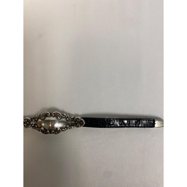 Op de afbeelding: Een zilverkleurige gesp met een sierlijk, ovaal ontwerp is bevestigd aan een zwarte, getextureerde riem. De gesp heeft een decoratieve rand en een gepolijst midden. De riem is gemaakt van een donker, mogelijk lederachtig materiaal.