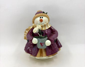 May include: A ceramic snowman figurine wearing a purple coat, hat, and scarf with gold stripes. The snowman holds a gray pot with white flowers and a red heart. The figurine has a carrot nose and black dot eyes.