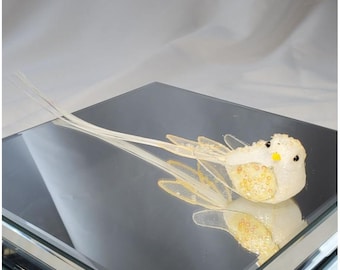 May include: A decorative bird ornament with a white body, yellow detailing, and long, delicate tail feathers. The bird has a textured surface and is on a mirrored surface. The wings are translucent with gold detailing.