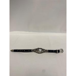 Op de afbeelding: Een zwarte leren riem met een zilverkleurige gesp en een decoratief zilverkleurig ovaal middelpunt. De riem heeft een textuurpatroon. Op een witte achtergrond.