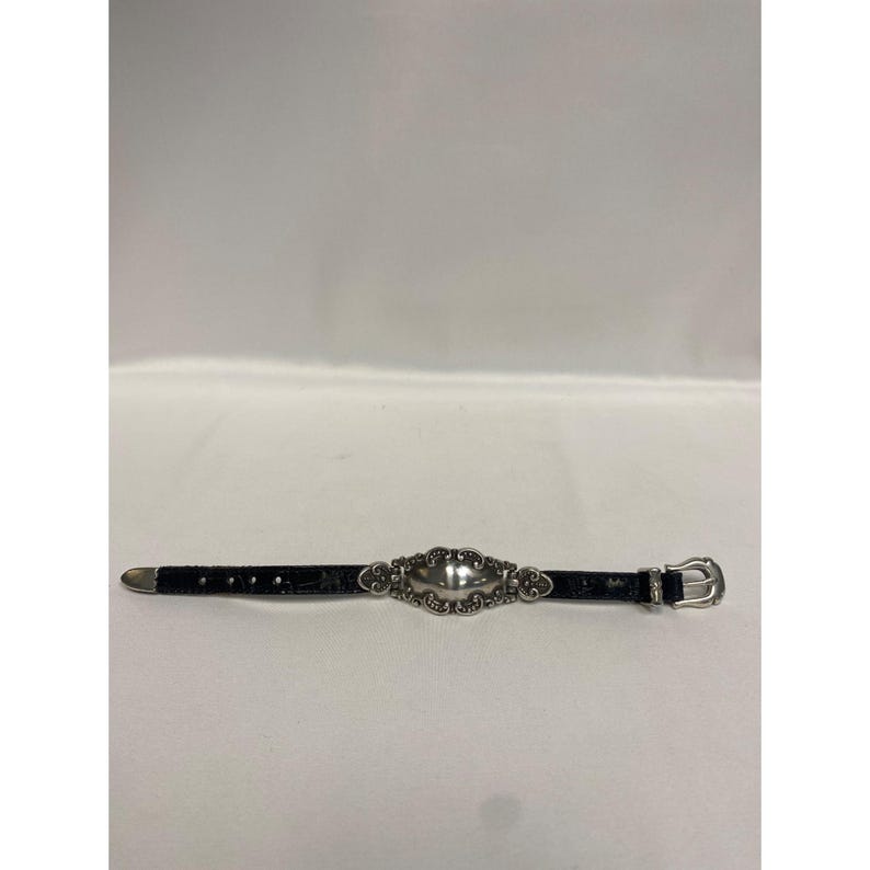 Op de afbeelding: Een zwarte leren riem met een zilverkleurige gesp en een decoratief zilverkleurig ovaal middelpunt. De riem heeft een textuurpatroon. Op een witte achtergrond.