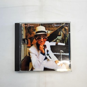 May include: CD case featuring Elton John's "Greatest Hits" album. The cover art shows Elton John in a white suit, pink sunglasses, and a white hat, seated near a piano. The CD case is black.
