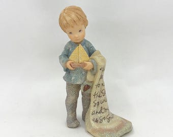 May include: A figurine of a young child holding a small yellow sailboat and a blanket. The child is wearing a blue shirt and glittery pants. The blanket has text and a heart design.
