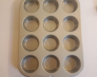 May include: A silver-colored, rectangular muffin tin with twelve circular cups. The tin has rounded edges and a small hole on one side. The brand name "Wilton" is printed on the top and bottom of the tin.