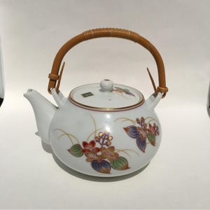 Pode incluir: Bule de cerâmica branco com uma alça tecida. O bule apresenta desenhos florais em vermelho, roxo e verde, acentuados com linhas douradas. A tampa tem um pequeno botão e uma borda dourada.