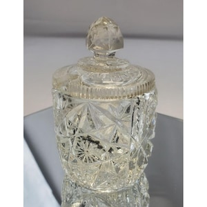 May include: A clear, faceted glass jar with a matching lid. The jar has a geometric pattern and a diamond-shaped finial on the lid. The jar is likely used for storing small items or as a decorative piece.