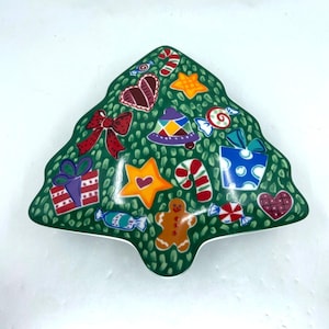 Puede incluir: Un plato de cerámica con forma de árbol de Navidad con fondo verde y decoraciones festivas. El plato presenta ilustraciones coloridas de regalos, bastones de caramelo, hombres de jengibre y corazones, perfecto para dulces navideños.