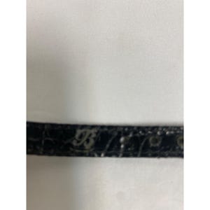 Op de afbeelding: Een close-up van een zwarte leren riem met een textuurpatroon. De riem heeft een zilveren gesp met de letters "B" en "93" in reli&euml;f. De achtergrond is een witte stof.