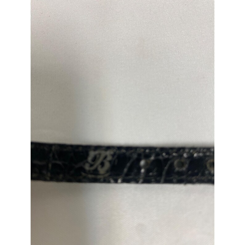 Op de afbeelding: Een close-up van een zwarte leren riem met een textuurpatroon. De riem heeft een zilveren gesp met de letters "B" en "93" in reli&euml;f. De achtergrond is een witte stof.