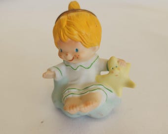 May include: A ceramic figurine of a child with yellow hair and blue eyes, wearing a white nightgown with green trim, sitting on a blue cloud. The child holds a yellow star. The figurine has freckles and a smiling expression.