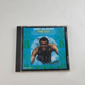 Puede incluir: Una caja de CD con el álbum "Pure Gold" de Harry Belafonte. La portada tiene un fondo turquesa con el nombre del artista y el título del álbum en blanco. La imagen muestra a Belafonte en el agua. El texto "Original Album Art" está en la parte inferior.