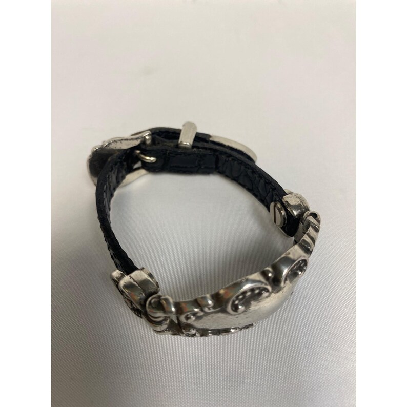 Op de afbeelding: Een zilverkleurige metalen en zwarte leren armband. De armband heeft een decoratieve zilveren sluiting en gesp. De zwarte leren band is vastgezet met zilveren klinknagels. De armband is ontworpen met een decoratieve zilveren metalen band.