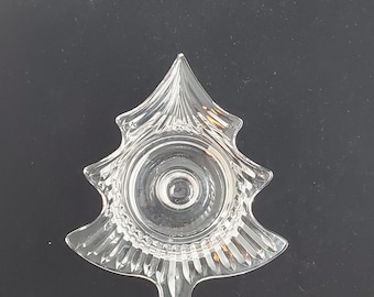 May include: A clear glass Christmas tree-shaped dish. The dish has a central circular indentation and is decorated with vertical and curved lines. The tree shape has a pointed top and a flared base. The dish is likely used for serving or decoration.