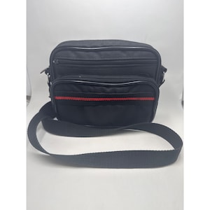 May include: Black messenger bag with a red stripe detail. The bag has a rectangular shape with multiple zippered compartments and a long, adjustable shoulder strap. The bag is made of a durable material and is suitable for everyday use.