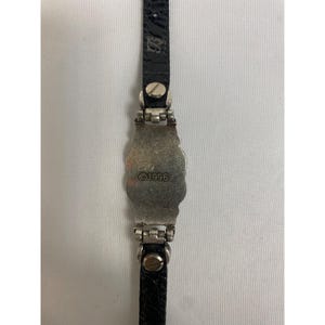 Op de afbeelding: Een zilverkleurige armband met een zwarte leren band. Het middelpunt van de armband heeft een sierlijk ontwerp en de tekst "&copy;1996". De leren band is bevestigd met zilverkleurige metalen hardware.