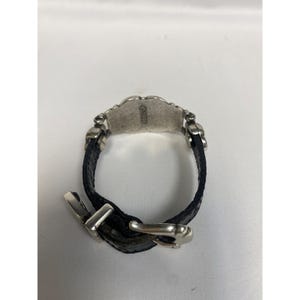 Op de afbeelding: Een zwart leren armband met een zilverkleurig decoratief middelpunt en een gesp. Het middelpunt heeft een geschulpte rand en de inscriptie "&copy;1998". De armband is ontworpen in een klassieke stijl.