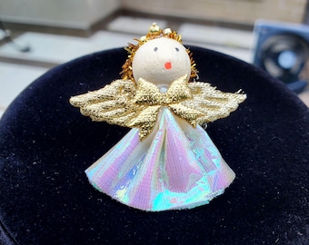 May include: A small angel figurine with a cream-colored head, gold wings, and a shimmering iridescent skirt. The angel has a gold halo and a bow at the chest. The figurine is likely a Christmas ornament or decorative item.