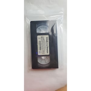 May include: A black VHS tape labeled "Grizzly & the Treasure" and "GOODTIMES" inside a clear plastic bag. The tape has white labels with black text. The tape is rectangular with two circular reels visible.
