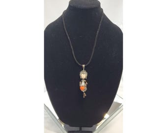 May include: A silver pendant necklace with a black cord. The pendant has a stylized figure with a cream face, orange torso, and silver details. The necklace is displayed on a black mannequin.